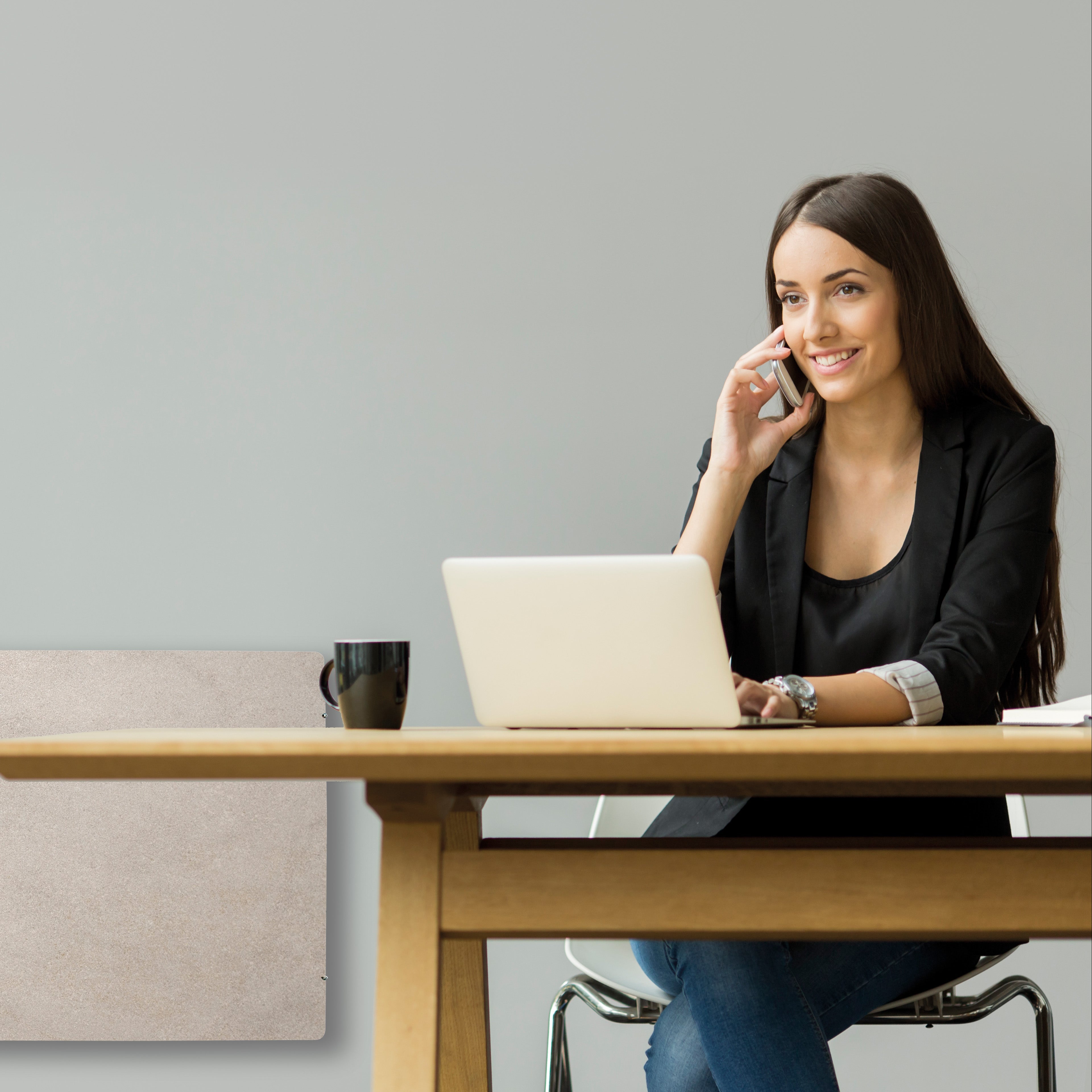 Junge Frau sitzt am Schreibtisch mit Laptop, spricht lächelnd am Telefon und arbeitet in moderner Büro-Umgebung.