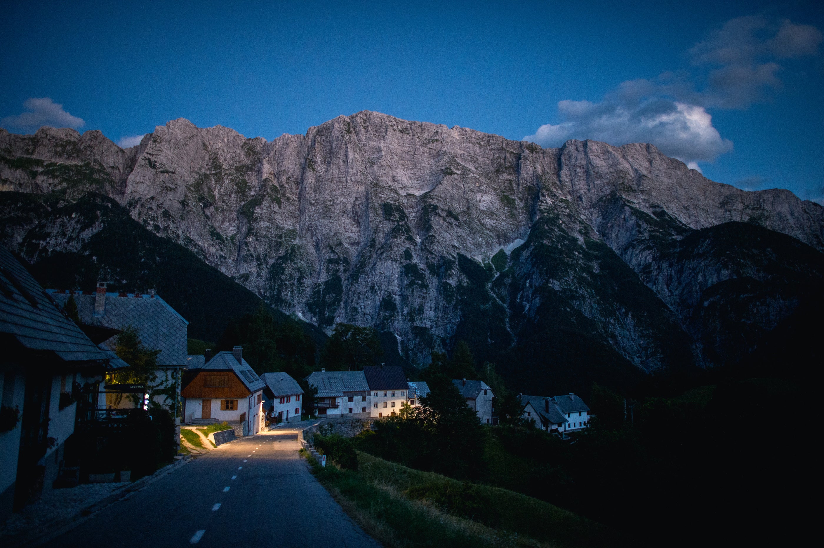 Bergdorf bei Sonnenuntergang mit alpiner Felswand im Hintergrund und traditionellen Häusern entlang einer kurvigen Strasse